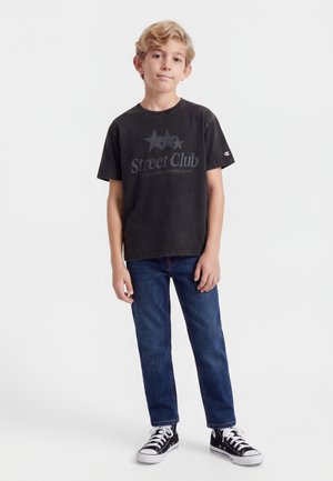 Garçon debout, portant un t-shirt noir "Street Club", un jean bleu et des baskets montantes noires, sur un fond blanc uni.