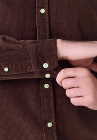 Brązowa koszula z korduru o teksturowanej tkaninie, z jas green guzikami i mankietami zapinanymi na dwa guziki. Jedna ręka zapina guzik mankietu.