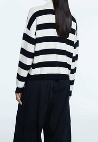 Personne aux longs cheveux foncés portant un pull à rayures horizontales noir et blanc et un pantalon noir ample, vue de dos sur un fond clair.