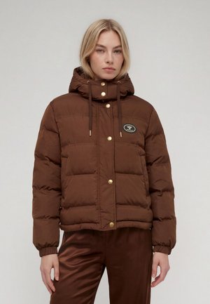 Femme portant une doudoune marron avec des boutons dorés et une capuche, debout devant un fond clair uni.