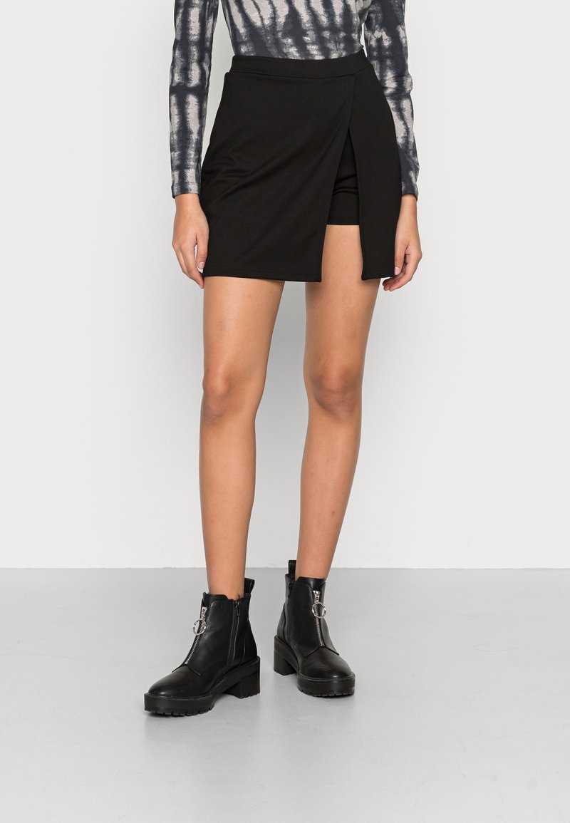Femme portant une jupe portefeuille asymétrique noire avec short intégré, un haut à manches longues teint en tie-dye, et des bottines noires à talons carrés.