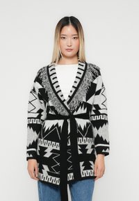 Ung kvinne iført en svart og hvit mønstret belted cardigan med frynsedetaljer over en hvit topp og blå jeans, som står mot en ensfarget bakgrunn.