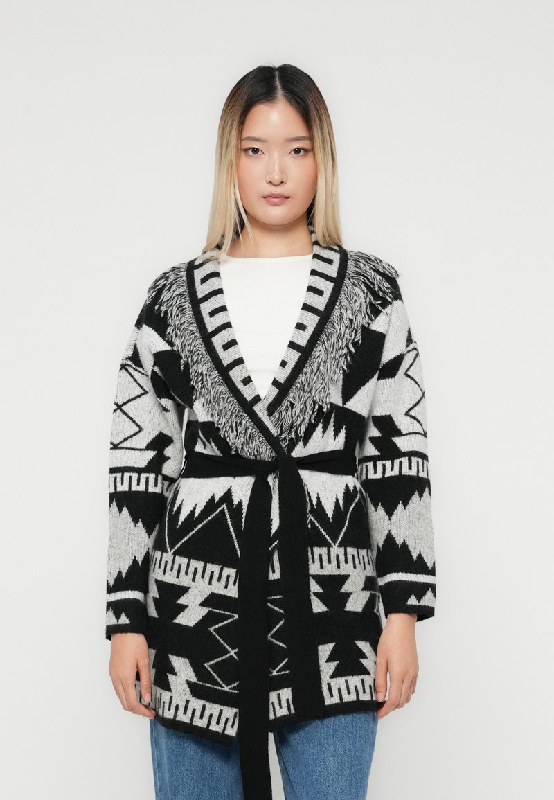 Ung kvinne iført en svart og hvit mønstret belted cardigan med frynsedetaljer over en hvit topp og blå jeans, som står mot en ensfarget bakgrunn.