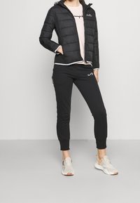 Svart puffjacka med huva, rosa t-shirt med logotyp, och svarta smala joggers. Lätta skor med mesh och accentfärger.