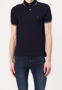 Polo bleu marine avec un col à rayures rouges et blanches, patte de boutonnage à deux boutons et petit logo sur la poitrine ; associé à un jean foncé.