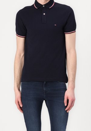 Polo bleu marine avec un col à rayures rouges et blanches, patte de boutonnage à deux boutons et petit logo sur la poitrine ; associé à un jean foncé.