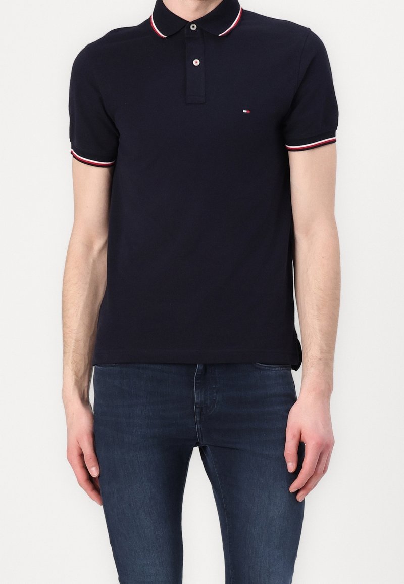 Polo bleu marine avec un col à rayures rouges et blanches, patte de boutonnage à deux boutons et petit logo sur la poitrine ; associé à un jean foncé.