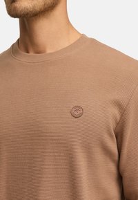 Primer plano de un hombre que lleva un suéter de canalé beige con un parche de cuero redondo en el pecho que dice "Est. 1993 Jeansspirit."