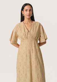 Beige klänning med V-ringning, korta volangärmar och broderade utskärningsmönster. Tyget har en texturerad yta med ett diamantmönster.
