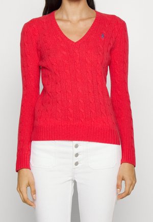 Kobieta ubrana w czerwony sweter z dzianiny z dekoltem w serek i małym niebieskim haftowanym logo na piersiach oraz białe jeansy z guzikami na froncie.