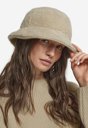 Beige bucket-hatt av texturerat, fluffigt material; har en bred brätte och subtil sömnad längs kanten.
