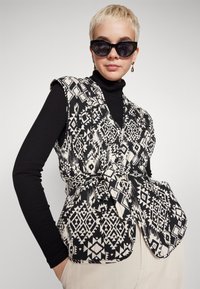 Gilet a fantasia in bianco e nero con vita cintata, realizzato in un tessuto leggero. Indossato sopra un dolcevita nero, con ampie aperture per le braccia.