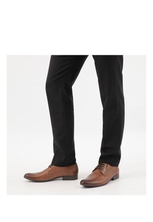 Homme portant des chaussures habillées en cuir marron associées à un pantalon formel noir, debout sur un fond blanc.
