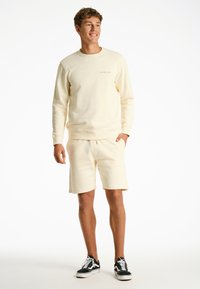 Felpa beige chiaro e pantaloni corti abbinati realizzati in morbido tessuto con una texture a coste. La felpa presenta un branding minimale sul petto.