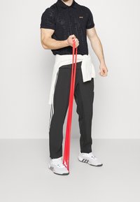 Black polo shirt with a grid pattern and embroidered logo, gray athletic pants with white stripes, holding red resistance bands, white sneakers.