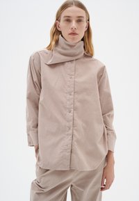 Lys beige kordfløjlsskjorte med høj krave, knapper fortil og lange ærmer. Har en afslappet pasform og tekstureret stof.