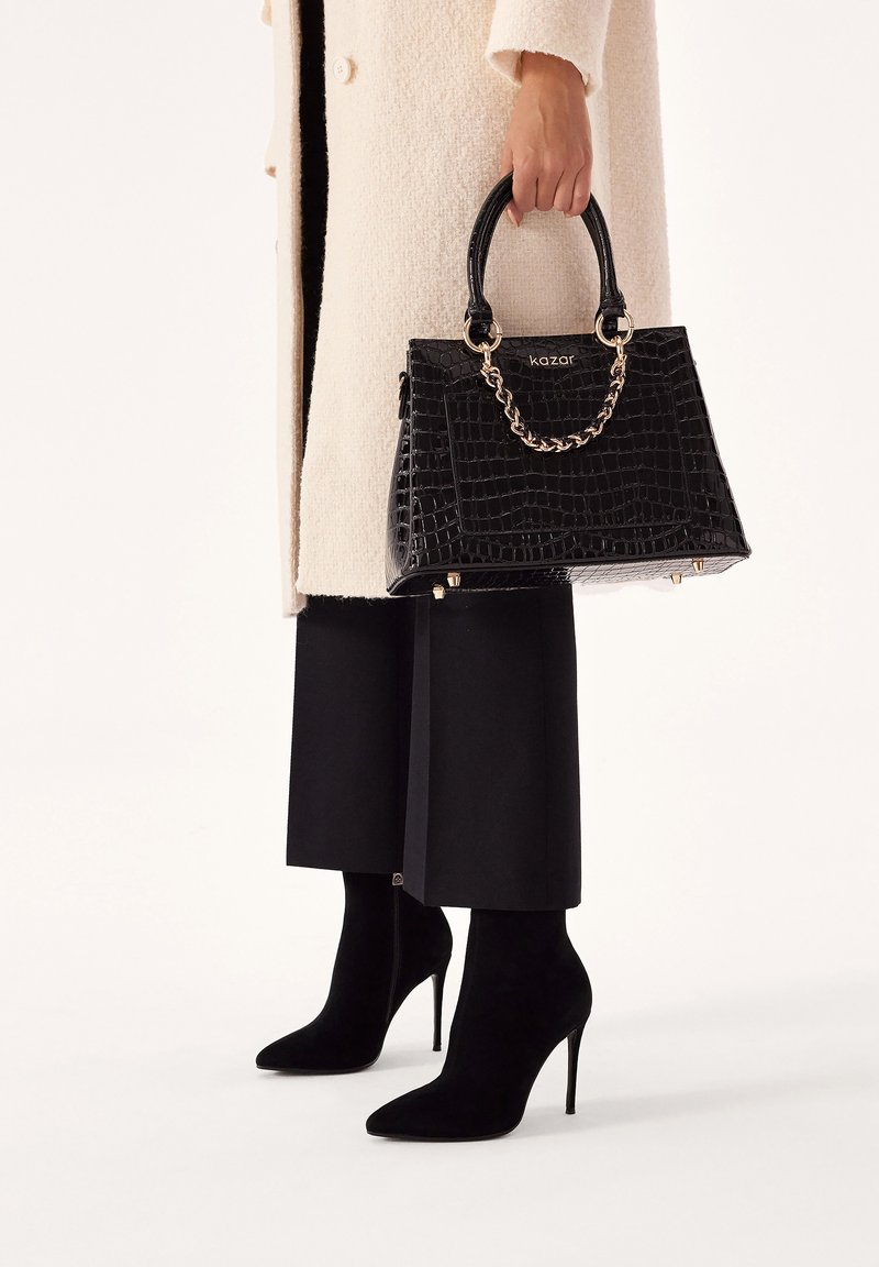 Black crocodile-embossed handbag with gold chain accents and structured design; worn with black wide-leg trousers and high-heeled black boots.