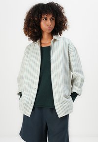 Kvinne med krøllete hår iført en oversized kremfarget skjorte med tynne blå striper over en mørkegrønn topp og marineblå shorts, hender i lommer.