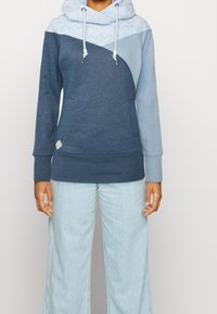 Felpa con cappuccio realizzata con un mix di tessuti blu scuro e chiaro, un design triangolare, parte superiore testurizzata e lacci. Abbinata a jeans chiari.