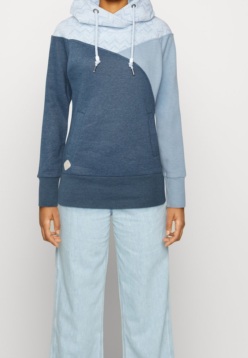 Felpa con cappuccio realizzata con un mix di tessuti blu scuro e chiaro, un design triangolare, parte superiore testurizzata e lacci. Abbinata a jeans chiari.