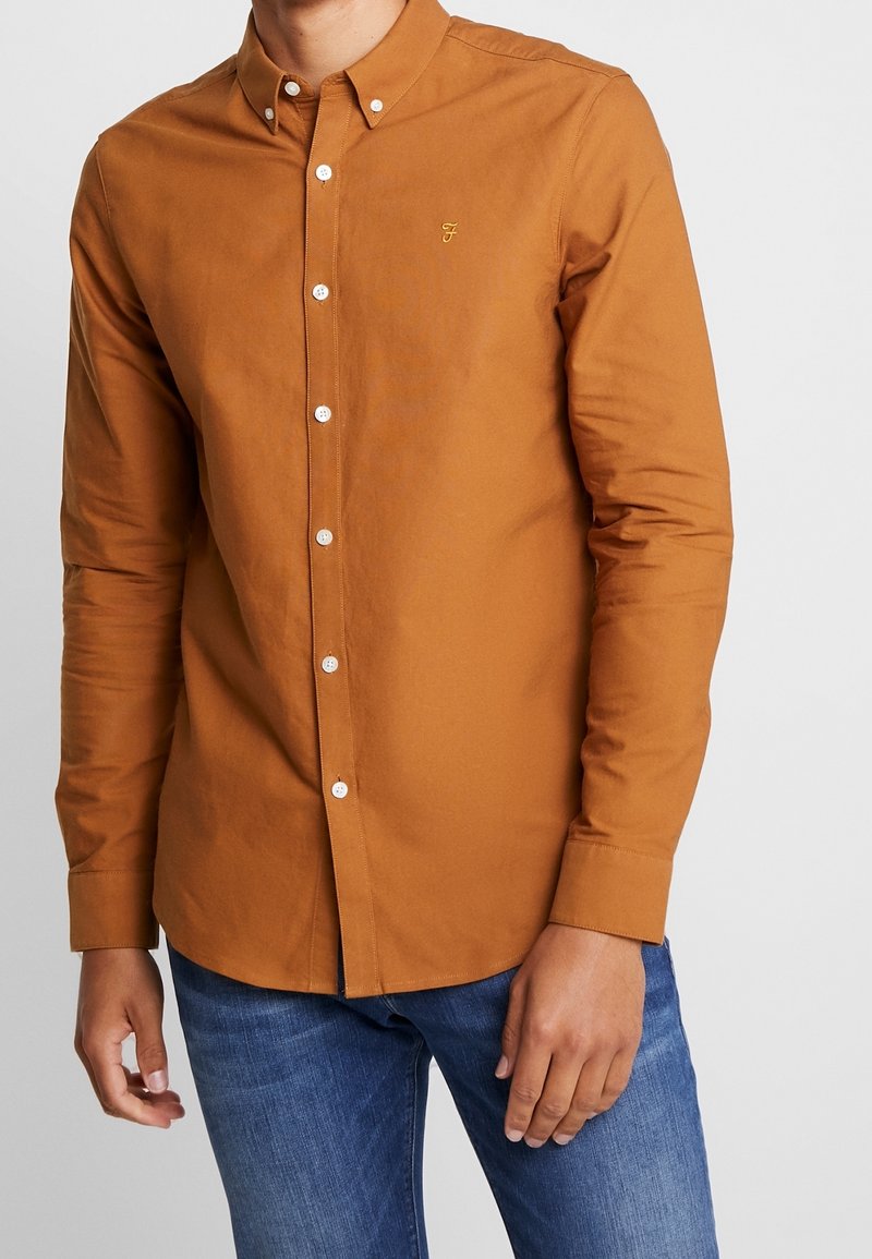 Lange mouwen button-up shirt in gebrand oranje katoen, voorzien van een subtiele logo, witte knopen en een klassieke kraag, gecombineerd met blauwe jeans.