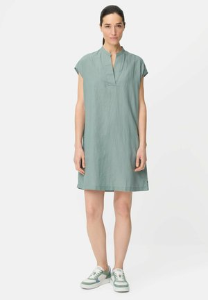 Kobieta stojąca przodem, ubrana w jasnoróżową sukienkę z krótkim rękawem oraz pasujące zielono-białe trampki na białym tle.