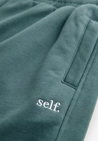 Teal värvi puuvillased joggers'id küljetasku ja valge tikandiga tekstiga "self." alumisel säärsel. Pehme tekstuur ja elastne vöökoht.