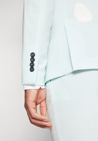 Svetle modrý blazer, rukáv se čtyřmi černými knoflíky, přiléhavý střih a hladká tkanina; bílé manžety košile viditelné pod ním.
