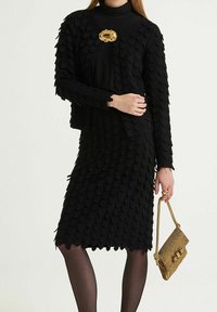 Ensemble robe noire à plusieurs couches et veste, en tissu texturé avec des détails festonnés. Broche dorée et sac à main doré complètent le look.