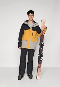 Geel-grijze sneeuwjas met zwarte accenten, gecombineerd met zwarte sneeuwbroek. Persoon houdt een ski met bindingen vast en draagt een skibril.