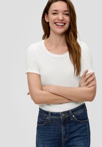 T-shirt blanc à manches courtes texturé avec un col rond, associé à un jean en denim bleu foncé avec des poches traditionnelles et des coutures.