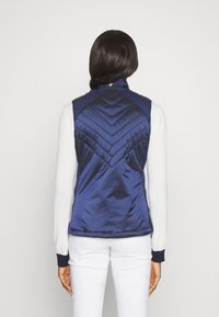 Námorně modrá, prošívaná pufovaná vesta s vysokým límcem a šikmým prošitím. Tkanina vypadá leskle, s kontrastním námořnickým lemováním kolem průramků.