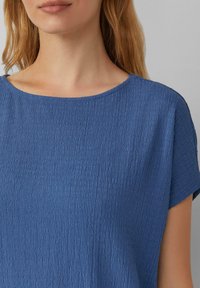 Blauw kortemouwtop met een gestructureerd oppervlak, heeft een wijde halslijn en een losse pasvorm, benadrukt het gekreukte patroon overal.