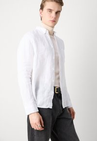 Weißes Leinenhemd mit Button-Down-Kragen, langen Ärmeln und lässiger Passform, kombiniert mit grauen Jeans und einem schwarzen Gürtel. Glatte Textur.