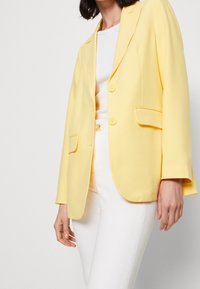 Żółta marynarka wykonana z gładkiego materiału, z wyciętymi klapami, dwoma przednimi kieszeniami i dwoma guzikami, zestawiona z białymi spodniami i bluzką.
