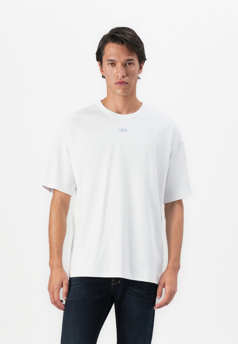 Young man wearing a plain white oversized t-shirt with a small blue logo center chest, paired with dark blue jeans, standing against white background.
