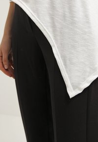 Weißes, strukturiertes Oberteil in einem gespaltenen Design, kombiniert mit schwarzen, maßgeschneiderten Hosen. Fokus auf die Stoffdetails und den Kontrast zwischen den Farben.