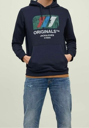 Mężczyzna w granatowej bluzie z kapturem z wielokolorowym logo "ORIGINALS™ JACK&JONES ©1990" i niebieskich, przetartych dżinsach, ręce w kieszeni bluzy.