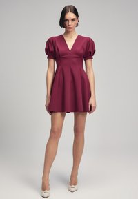 Burgundské mini šaty s nabíranými rukávy, hlubokým V-výstřihem a skládací sukní, vyrobené z hladké, strukturované tkaniny. Doplňují je elegantní lodičky.
