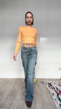 Άτομο που περπατάει σε εσωτερικό χώρο φορώντας πορτοκαλί κοντή μπλούζα με μακριά μανίκια και μπλε τζιν, με ένα τατουάζ ορατό στην κάτω κοιλιακή περιοχή.