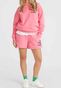 Rosa Kapuzenpullover mit Grafiktext, kombiniert mit passenden rosa Shorts mit Druck. Weißes T-Shirt darunter, grüne Socken und weiße Turnschuhe.