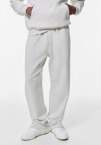 Persoon in lichtgrijze sweatpants, witte sneakers en een bijpassende lichtgrijze sweatshirt met handen in de zakken.