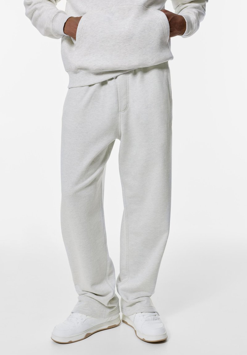 Persoon in lichtgrijze sweatpants, witte sneakers en een bijpassende lichtgrijze sweatshirt met handen in de zakken.