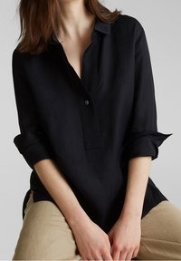 Frau trägt ein schwarzes Langarmshirt mit hochgekrempelten Ärmeln und beigen Hosen, sitzt mit den Händen auf dem Schoß.