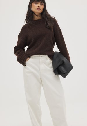 Vrouw met een donkergroene, zachte trui en witte broek met hoge taille, die een zwart jasje in één hand vasthoudt tegen een effen achtergrond.