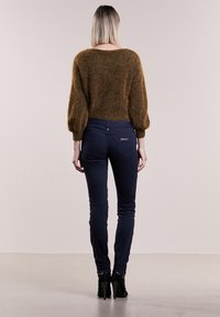 Brązowy, futrzasty sweter z opadającymi rękawami w kształcie balonu, w połączeniu z obcisłymi, ciemnoniebieskimi dżinsami z tylnymi kieszeniami i detalem logo. Czarne buty na obcasie.