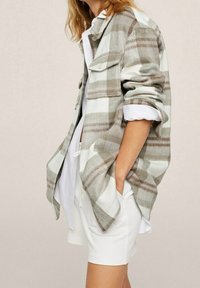 Camisa de cuadros en tonalidades beige y blanca, con bolsillos en el pecho, mangas enrolladas y un ajuste relajado, usada sobre una blusa blanca y pantalones cortos.