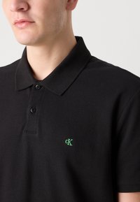 Vyras dėvi juodą polo marškinėlį su žaliu „cK“ logotipu kairėje krūtinės pusėje, matomas nuo kaklo iki vidurio liemens.