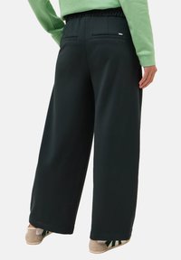 Pantaloni a gamba larga verde scuro con vita elasticizzata, dotati di due tasche posteriori e una texture liscia. Indossati con scarpe chiare.