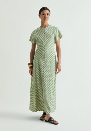 Una mujer está de pie vistiendo un vestido a cuadros verde y amarillo hasta el tobillo con mangas cortas, sandalias oscuras y tres pulseras en su muñeca derecha.